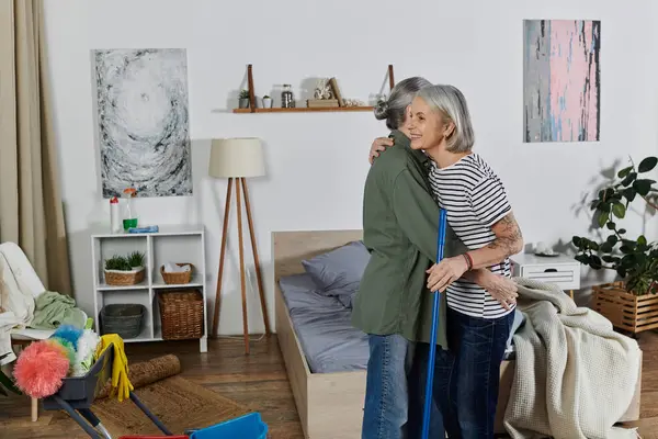 A mature lesbian couple takes a break from cleaning their modern apartment, sharing a warm embrace. — Stock Photo