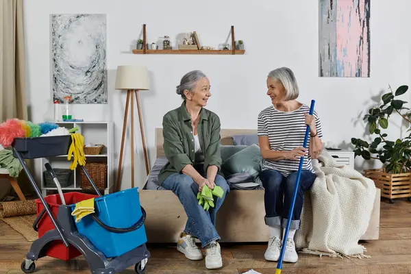 Duas mulheres lésbicas maduras em traje casual limpam seu apartamento moderno. Um senta-se na cama segurando luvas, o outro segura uma esfregona. — Fotografia de Stock
