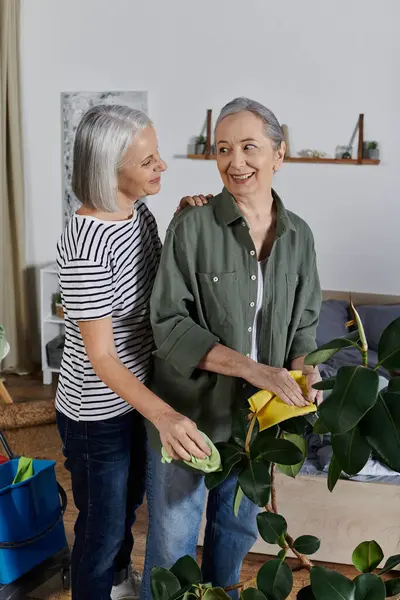 Two mature lesbian women in casual attire dust plants together in their modern apartment. — Stock Photo