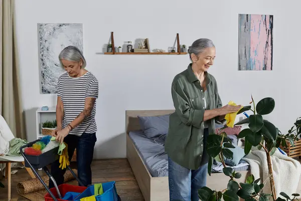 Two mature lesbian women in casual attire clean their modern apartment: one dusts a plant, the other sorts cleaning supplies. — Stock Photo