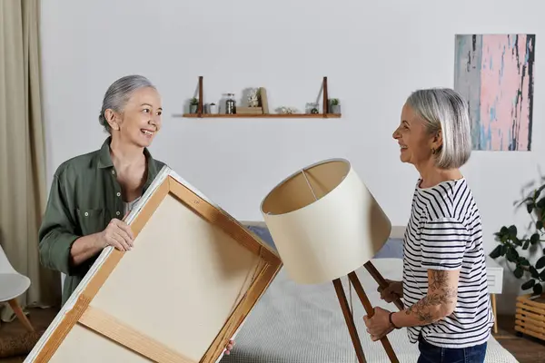Two mature women, dressed casually, are carrying furniture in their modern apartment. — Stock Photo
