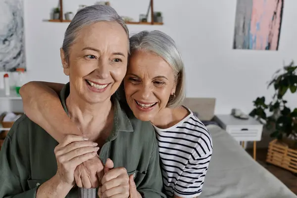 Two mature women embrace and laugh together in a modern apartment, showcasing a loving relationship. — Stock Photo