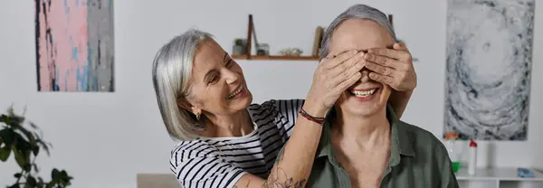 A mature lesbian couple in their modern apartment, one woman playfully covering her partners eyes. — Stock Photo