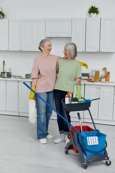 Two casual mature lesbians clean their modern apartment. Woman on left mops, her wife on right holds a cleaning cart with supplies. — Stock Photo