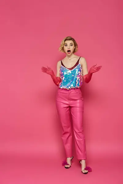 A woman in a pink outfit stands against a pink background, looking surprised. — Stock Photo