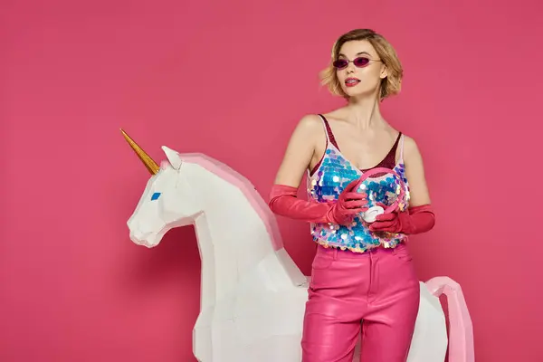 A woman in a bright pink outfit stands next to a white unicorn against a pink backdrop. — Stock Photo