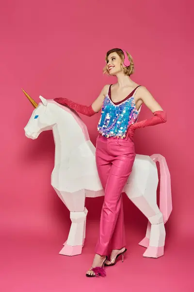 Woman in pink sequined top stands by white unicorn on pink backdrop. — Stock Photo