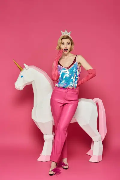 Una mujer con pantalones rosados, un top de lentejuelas y una corona posa frente a un unicornio blanco. - foto de stock