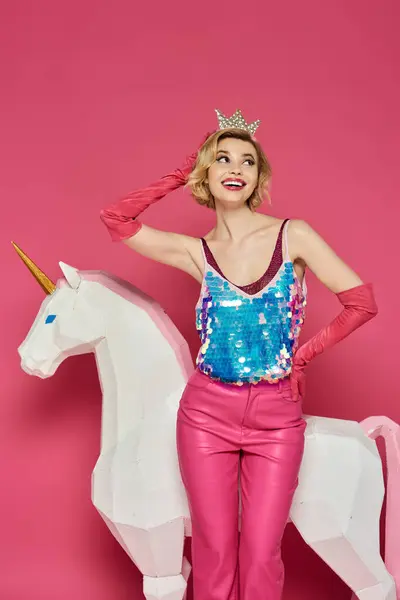 A woman in a sparkly top, pink pants, and a crown poses with a white unicorn. — Stock Photo