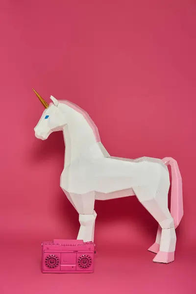 Un unicornio de origami blanco se levanta frente a un fondo rosa con una caja de resonancia rosa a sus pies. - foto de stock