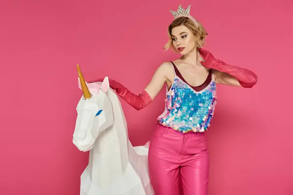Una mujer con una blusa brillante y pantalones rosas posa con un unicornio blanco sobre un fondo rosa. - foto de stock