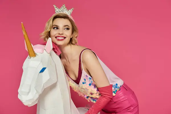 Mujer con corona y traje rosa posa con unicornio blanco sobre fondo rosa. - foto de stock