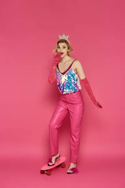 A woman in a pink outfit and crown stands on a pink skateboard against a pink background. — Stock Photo