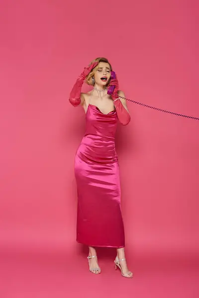 A woman in a pink satin dress and gloves uses a vintage phone against a pink background. — Stock Photo