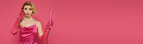 A woman in a pink dress and gloves holds a pink hair straightener against a solid pink background. — Stock Photo