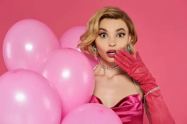 Uma mulher surpreso em um vestido rosa e luvas fica em meio a um aglomerado de balões cor de rosa. — Fotografia de Stock