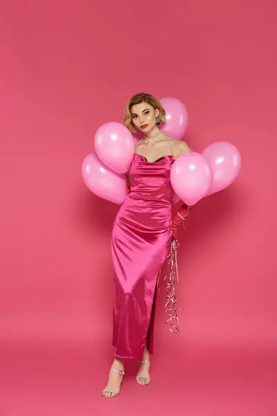 A woman in a vibrant pink dress poses with a cluster of pink balloons against a pink background. — Stock Photo