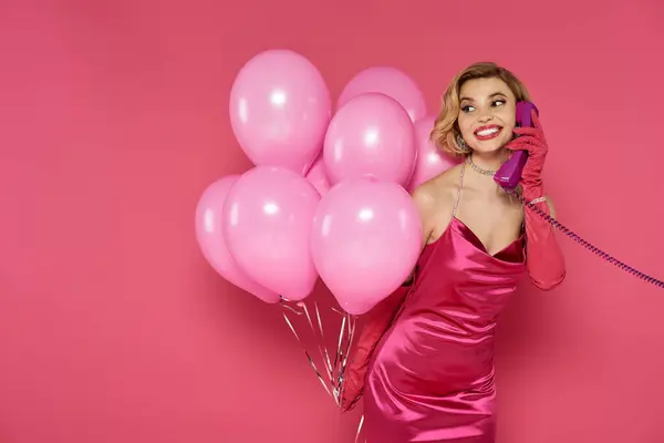 A woman in a vibrant pink dress holds a bunch of pink balloons and talks on a vintage phone. — Stock Photo