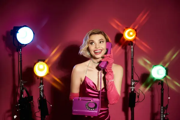 Mulher de vestido rosa e luvas sorri no telefone vintage em meio a luzes de palco. — Fotografia de Stock