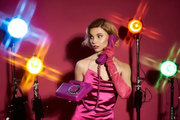 A woman in a pink dress and gloves speaks on a vintage phone while posing under studio lights. — Stock Photo