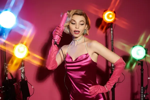 A woman in a pink satin dress and long gloves poses with a curling iron under bright studio lights. — Stock Photo