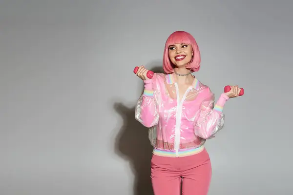 A woman with pink hair, wearing a pink see-through jacket, lifts weights against a gray backdrop. — Stock Photo