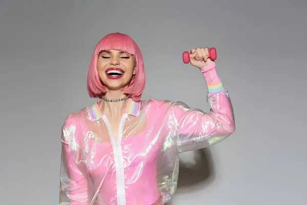 A woman in a pink wig and transparent jacket lifts a pink dumbbell with a joyous expression. — Stock Photo