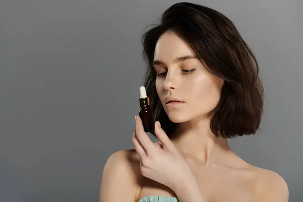 A woman with short brunette hair holds a bottle of serum, gazing at it with a serene expression. — Stock Photo