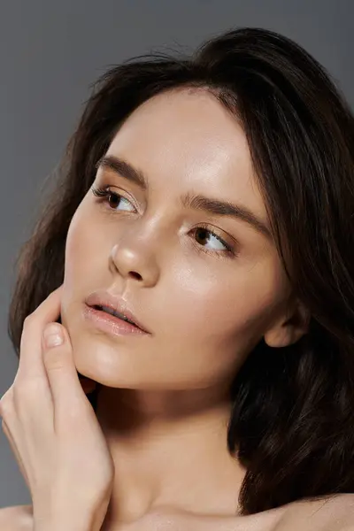 Woman with long dark hair poses against plain gray backdrop, hand delicately on cheek. — Stock Photo