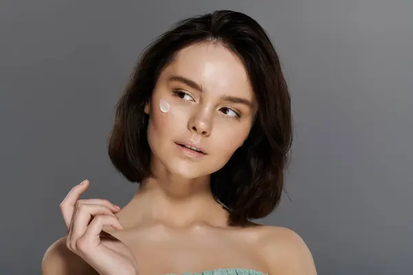 A young woman with short dark hair applies moisturizer to her cheek against a plain gray background. — Stock Photo