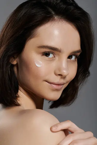 Una bella donna con i capelli corti scuri sorride delicatamente mentre applica la crema idratante sulla guancia. — Foto stock