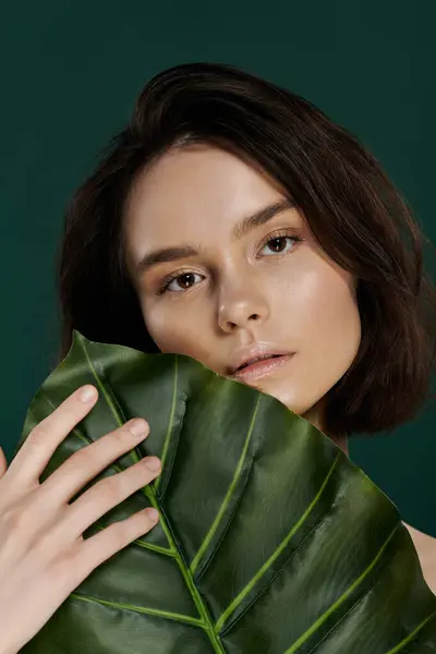 Woman with short brown hair poses against green backdrop, holding large green leaf. — Stock Photo
