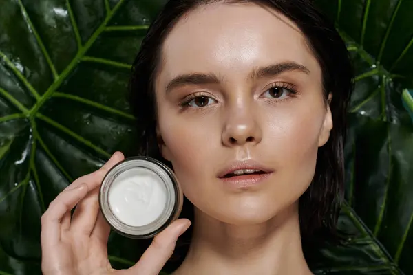 A woman with clear skin holds a container of cream in front of a vibrant green backdrop. — Stock Photo