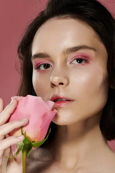 A woman with pink eyeshadow holds a soft pink rose against her lips. — Stock Photo