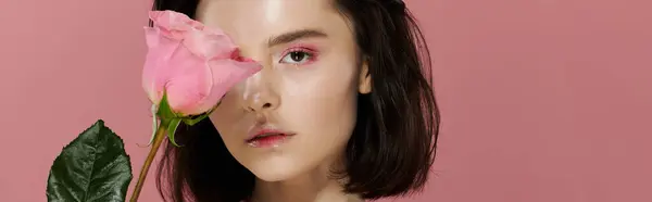 A woman with dark hair poses in front of a pink backdrop, a single pink rose obscuring one eye. — Stock Photo