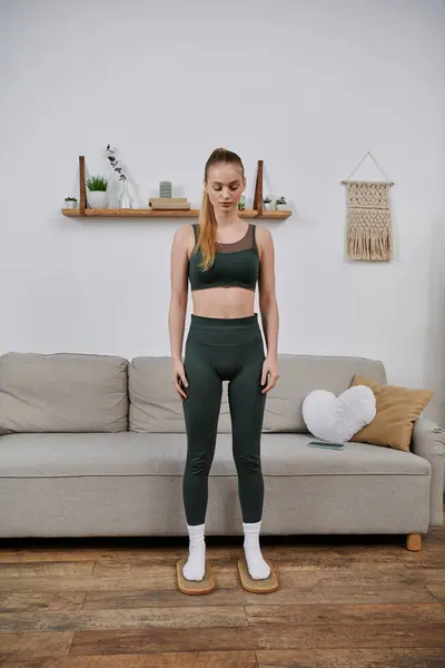A young woman in green athletic wear stands in a living room, her eyes closed as if lost in thought. — Stock Photo