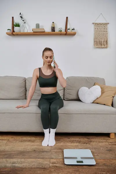 Donna con attrezzatura da allenamento sul divano che tiene il telefono, con scala di peso. — Foto stock