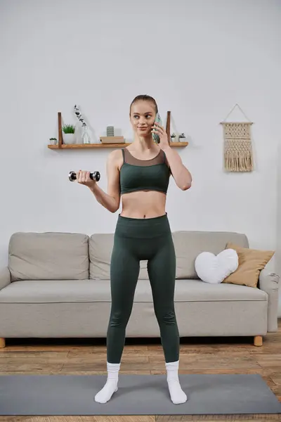 A young woman lifts weights while on a phone call in her living room. — Stock Photo