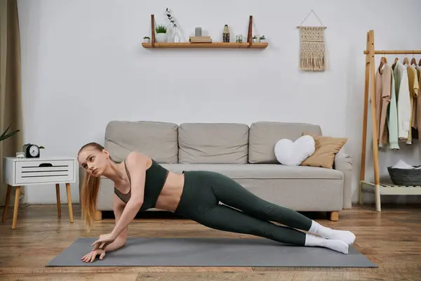 A young woman performs a side plank exercise on a yoga mat in her living room. — Stock Photo