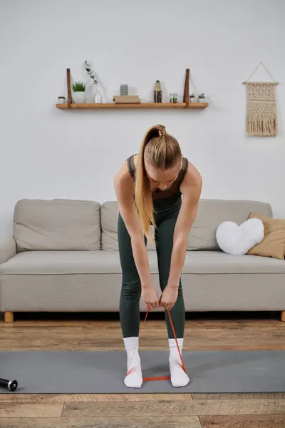 Donna in abiti da allenamento si estende con una banda di resistenza rossa su un tappetino yoga in soggiorno. — Foto stock