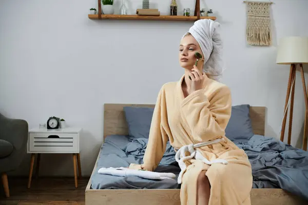 A young woman in a bathrobe sits on her bed, using a facial roller to pamper herself. — Stock Photo