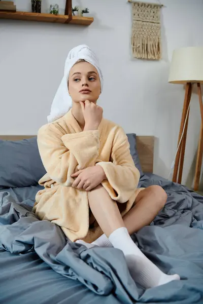A young woman in a bathrobe sits on a bed, thoughtfully resting her chin on her hand. — Stock Photo