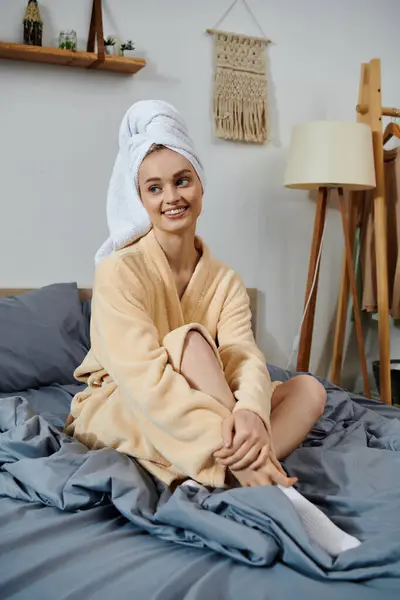 Una giovane donna in accappatoio si siede su un letto, sorridendo e distogliendo lo sguardo dalla telecamera. — Foto stock