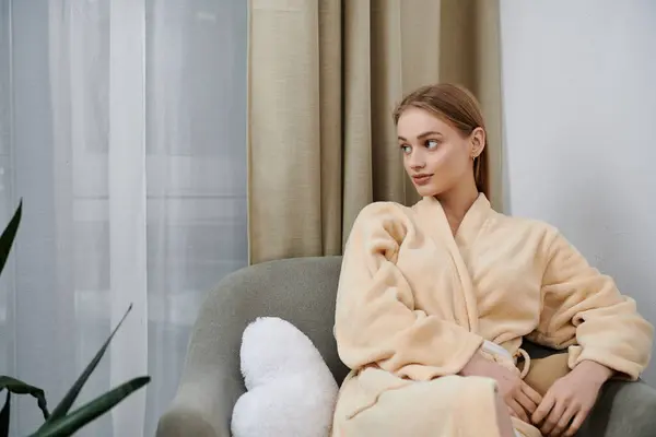 A young woman relaxes in a plush robe, seated on a cozy couch near a window. — Stock Photo