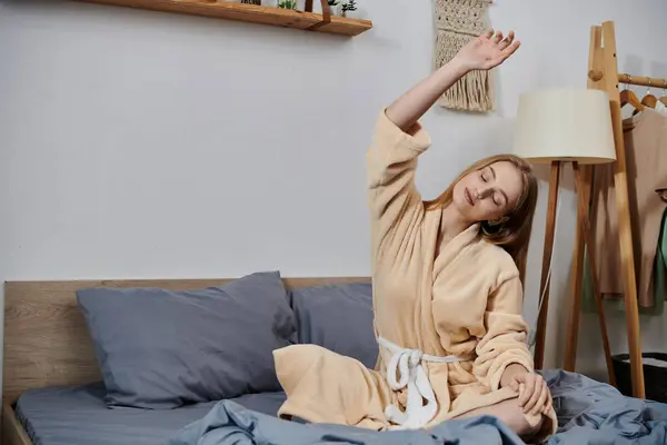 A young woman stretches in a robe on a bed at home. — Stock Photo