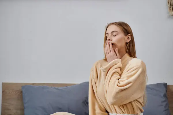 A young woman in a robe sits on her bed and yawns. — Stock Photo