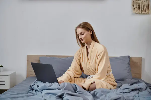 A young woman in a robe sits on a bed, working on a laptop. — Stock Photo