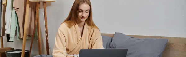 Una giovane donna che indossa un accappatoio utilizza un computer portatile mentre è seduta su un letto. — Foto stock
