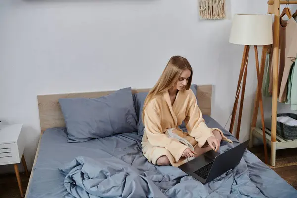 A young woman sits on her bed, wearing a robe, and uses her laptop. — Stock Photo