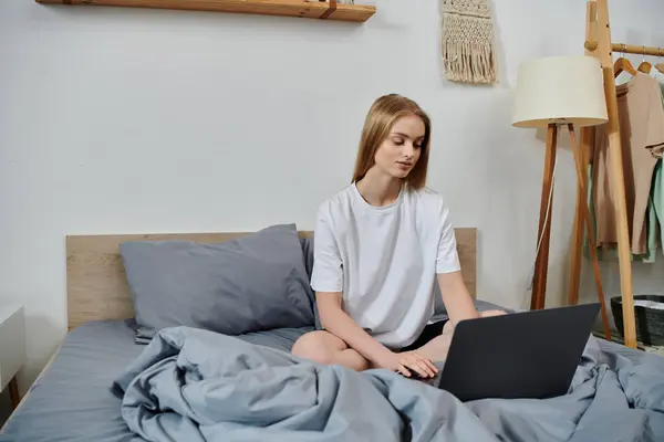 A young woman sits on a bed, working on a laptop. — Stock Photo